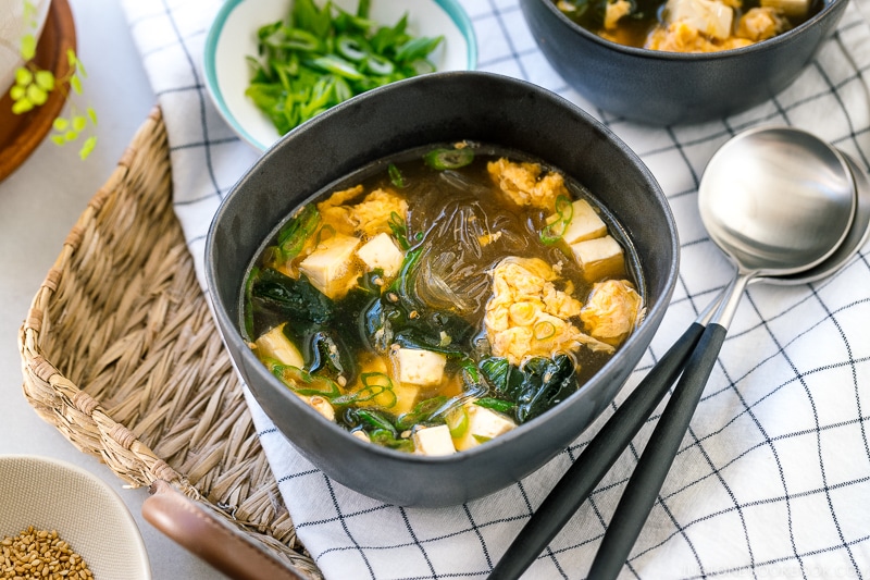 Black ceramic bowls containing glass noodle soup with tofu, fluffy egg, and wakame seaweed.