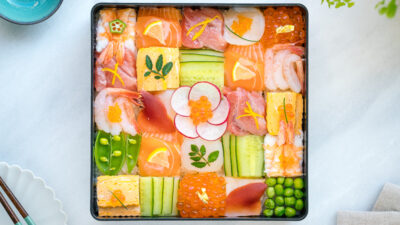 A Japanese lacquer box containing colorful Mosaic Sushi that's made of checkerboard pattern of various sashimi, tamago, and cucumber laid over sushi rice.