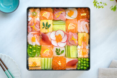 A Japanese lacquer box containing colorful Mosaic Sushi that's made of checkerboard pattern of various sashimi, tamago, and cucumber laid over sushi rice.
