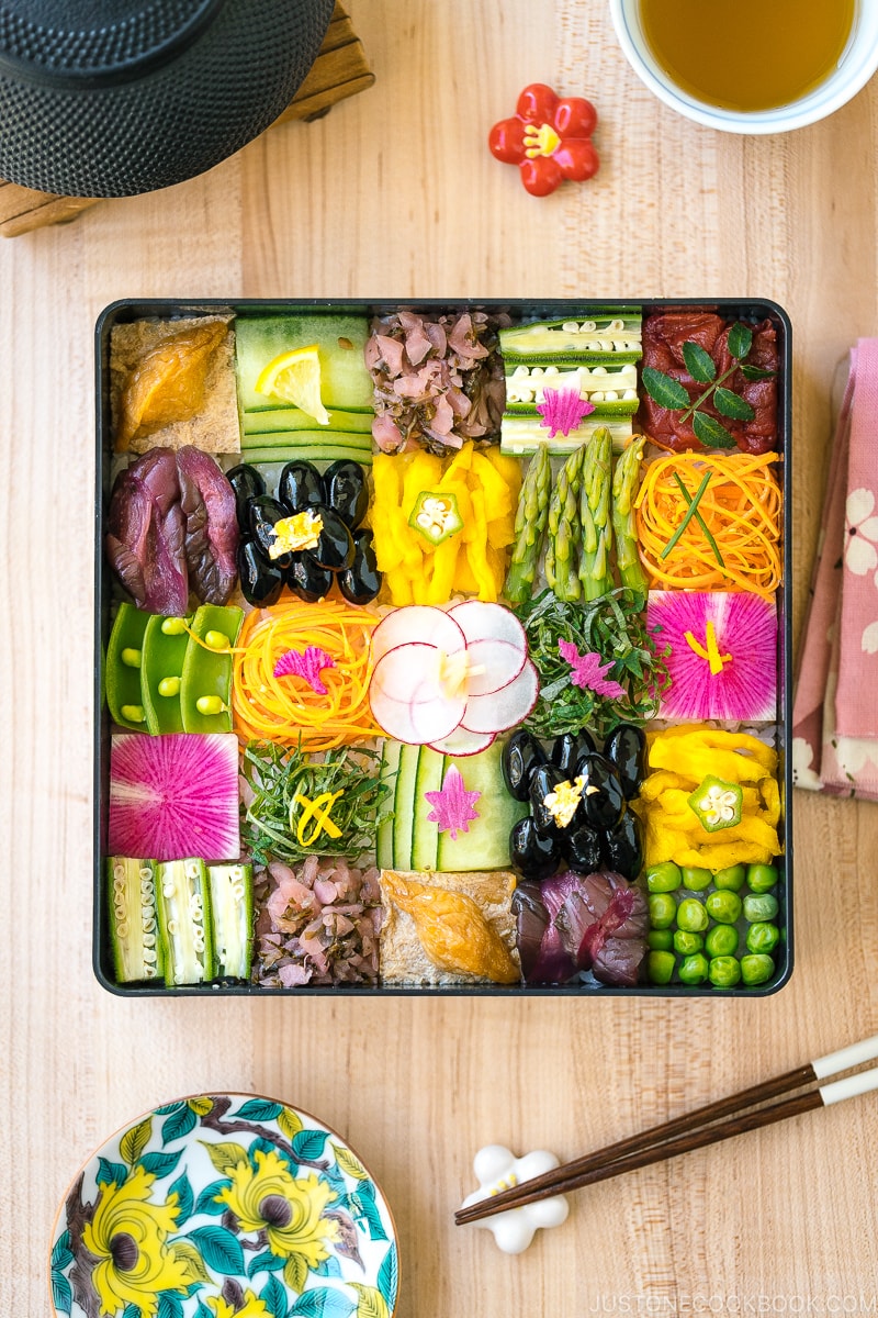 A Japanese lacquer box containing colorful Mosaic Sushi that's made of checkerboard pattern of various vegan-friendly ingredients laid over sushi rice.