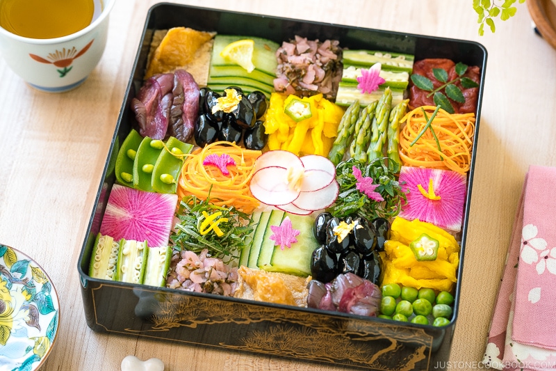 A Japanese lacquer box containing colorful Mosaic Sushi that's made of checkerboard pattern of various vegan-friendly ingredients laid over sushi rice.