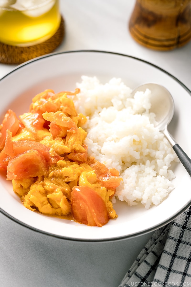 A white ceramic shallow bowl containing Stir-Fried Tomatoes and Eggs over steamed rice.