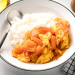 A white ceramic shallow bowl containing Stir-Fried Tomatoes and Eggs over steamed rice.