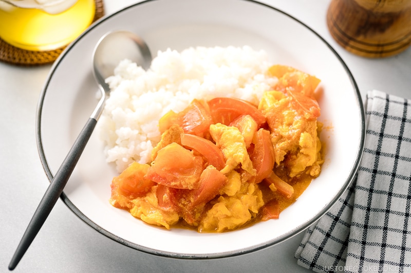 A white ceramic shallow bowl containing Stir-Fried Tomatoes and Eggs over steamed rice.