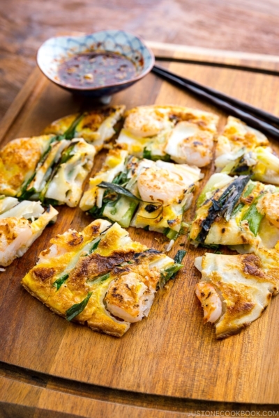 A white plate containing Korean Pancake (Pajeon) served with a dipping sauce.