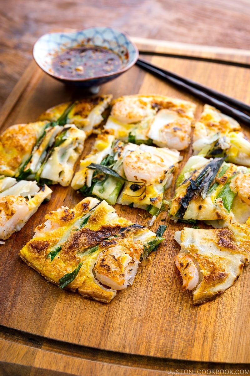 A white plate containing Korean Pancake (Pajeon) served with a dipping sauce.