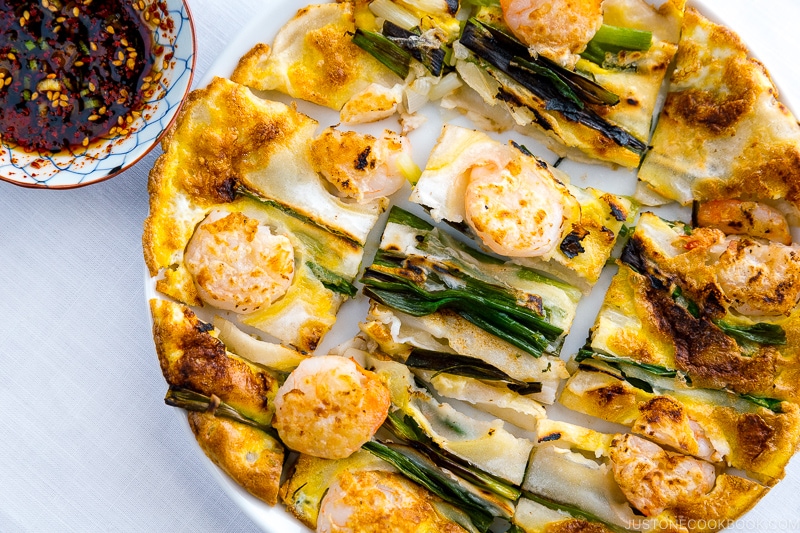 A white plate containing Korean Pancake (Pajeon) served with a dipping sauce.