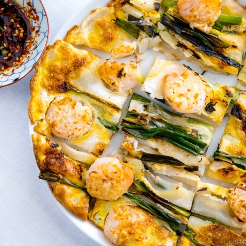 A white plate containing Korean Pancake (Pajeon) served with a dipping sauce.