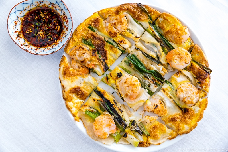 A white plate containing Korean Pancake (Pajeon) served with a dipping sauce.