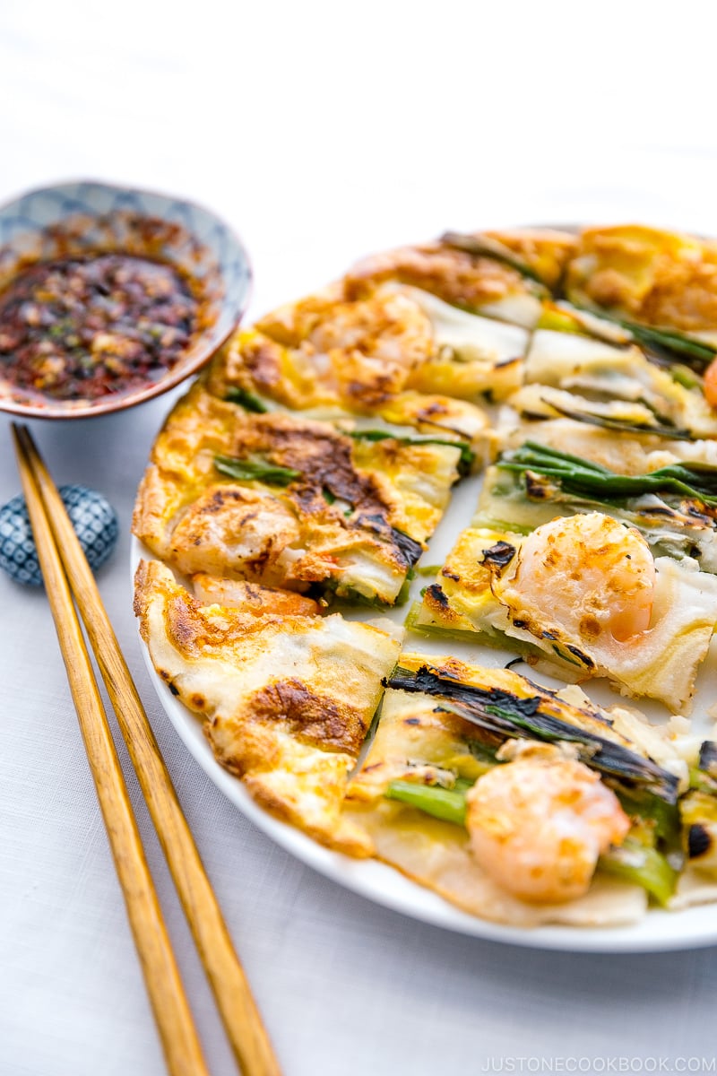 A white plate containing Korean Pancake (Pajeon) served with a dipping sauce.