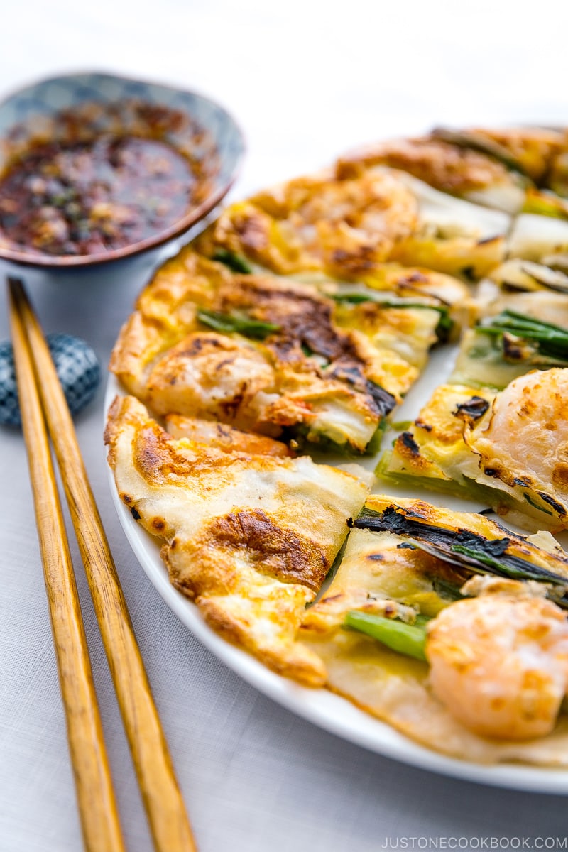 A white plate containing Korean Pancake (Pajeon) served with a dipping sauce.
