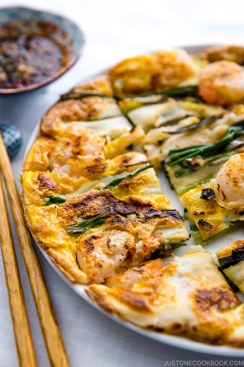 A white plate containing Korean Pancake (Pajeon) served with a dipping sauce.