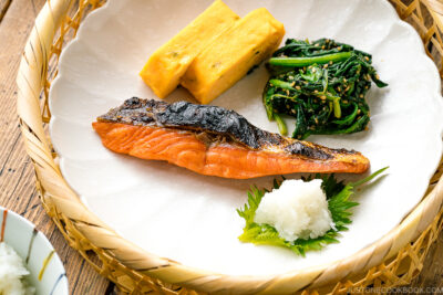A white plate containing Japanese salted salmon (Shiozake), tamagoyaki, and spinach salad.