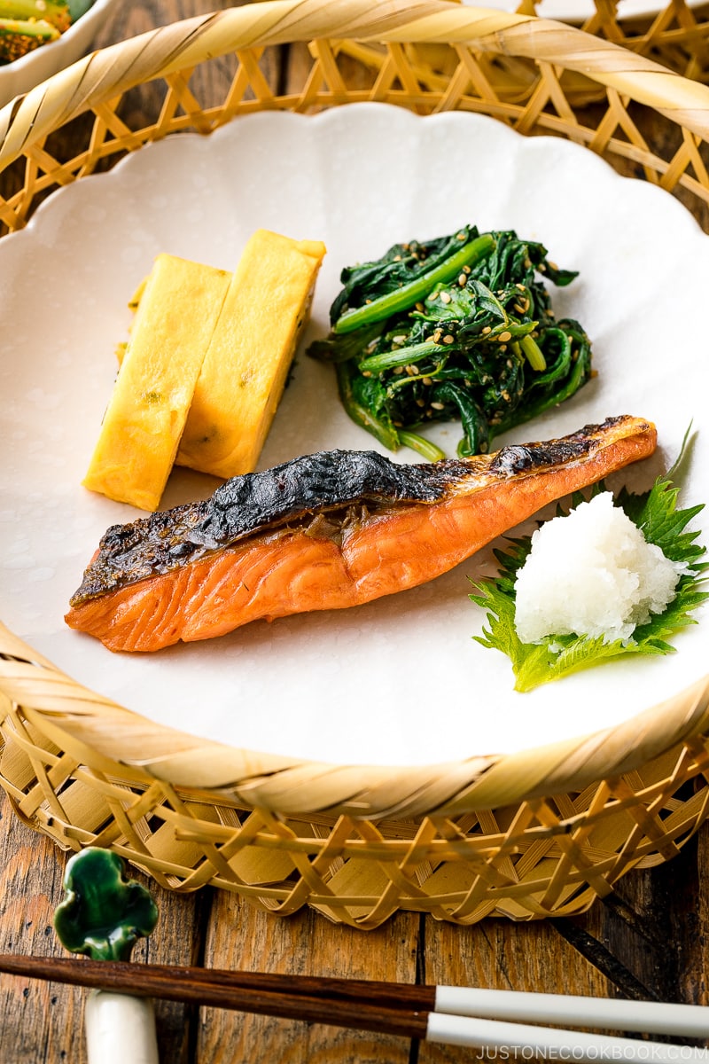 A white plate containing Japanese salted salmon (Shiozake), tamagoyaki, and spinach salad.