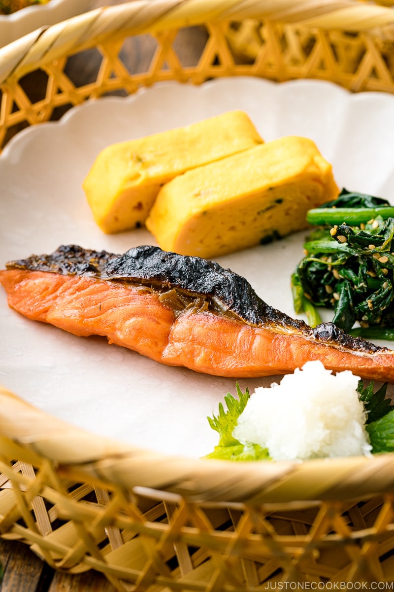 A white plate containing Japanese salted salmon (Shiozake), tamagoyaki, and spinach salad.