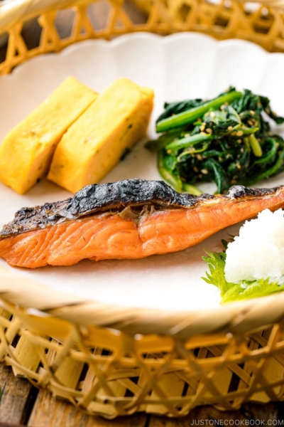 A white plate containing Japanese salted salmon (Shiozake), tamagoyaki, and spinach salad.