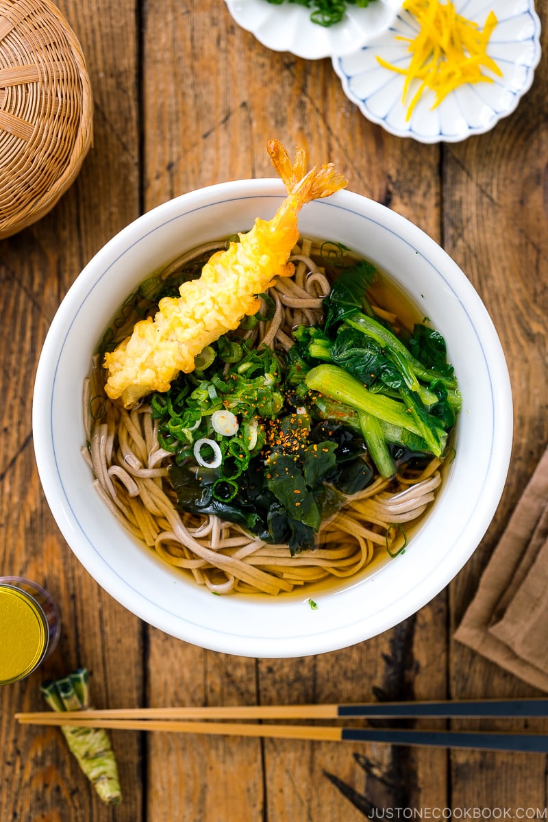 A bowl of soba noodle soup topped with shrimp tempura, wakame seaweed, blanched komatsuna, and green onions.