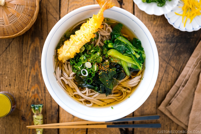 A bowl of soba noodle soup topped with shrimp tempura, wakame seaweed, blanched komatsuna, and green onions.