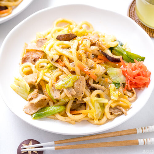 A white round plate containing Yaki Udon (Japanese Stir-Fried Udon Noodles) garnished with red pickled ginger.