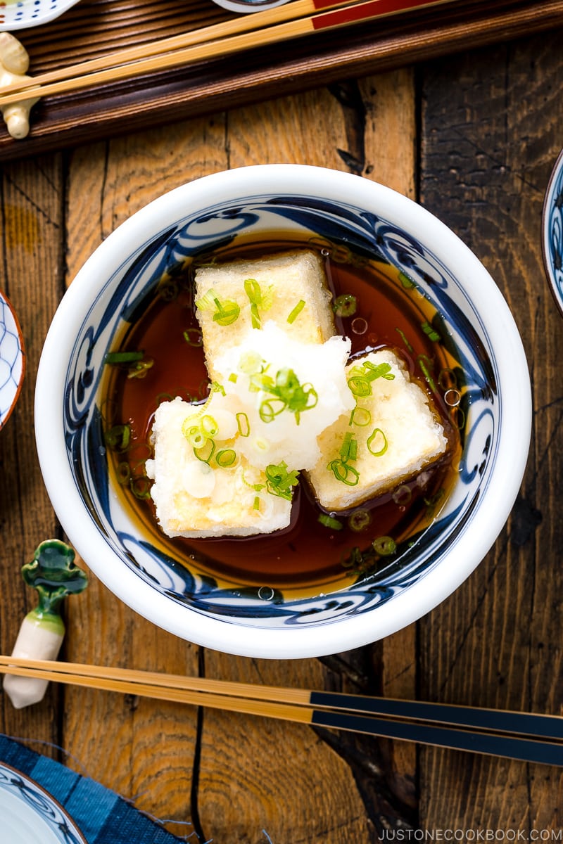 A ceramic bowl containing Agedashi Tofu (Agedashi Dofu) topped with grated daikon, chopped scallions, and grated ginger.