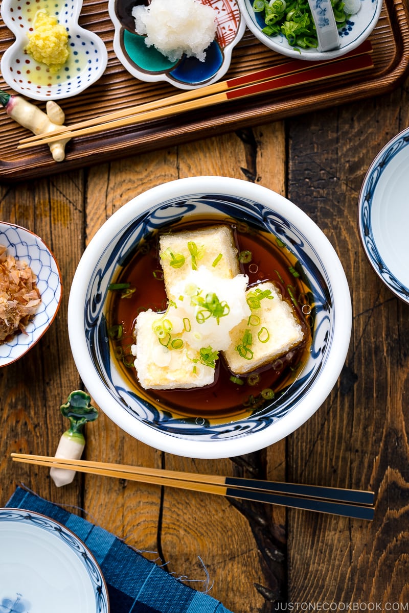 A ceramic bowl containing Agedashi Tofu (Agedashi Dofu) topped with grated daikon, chopped scallions, and grated ginger.