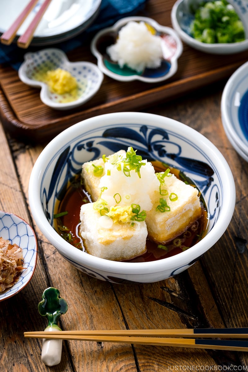 A ceramic bowl containing Agedashi Tofu (Agedashi Dofu) topped with grated daikon, chopped scallions, and grated ginger.