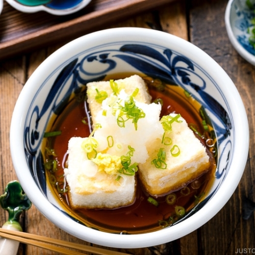 A ceramic bowl containing Agedashi Tofu (Agedashi Dofu) topped with grated daikon, chopped scallions, and grated ginger.