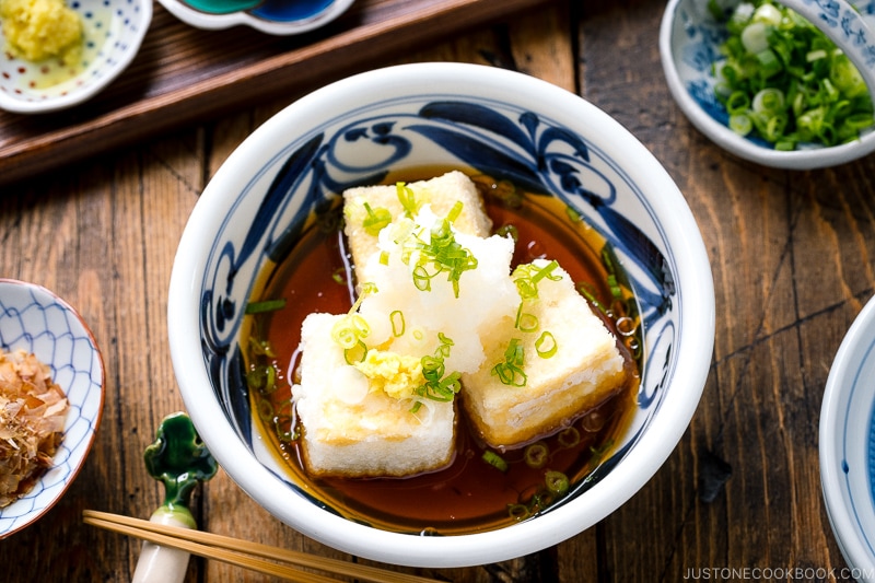 A ceramic bowl containing Agedashi Tofu (Agedashi Dofu) topped with grated daikon, chopped scallions, and grated ginger.