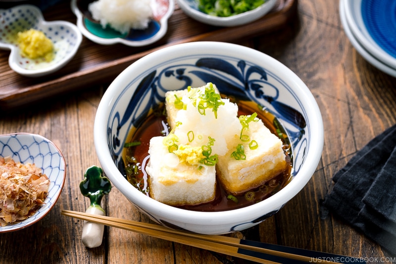 A ceramic bowl containing Agedashi Tofu (Agedashi Dofu) topped with grated daikon, chopped scallions, and grated ginger.
