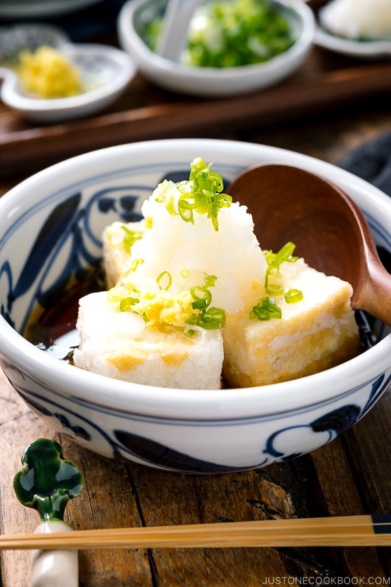 A ceramic bowl containing Agedashi Tofu (Agedashi Dofu) topped with grated daikon, chopped scallions, and grated ginger.