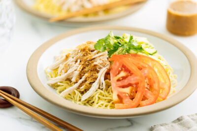 A ceramic plate containing Hiyashi Chuka (Cold Ramen) with Sesame Miso Sauce. Chilled noodles are served with sliced tomatoes, shredded chicken, and cucumber.
