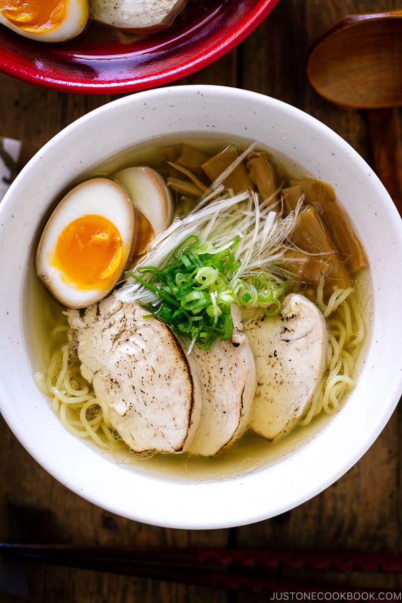 A white bowl containing Shio Ramen, topped with Chicken Chashu, Ramen Egg, bamboo shoot, white negi, and green onion.