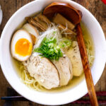 A white bowl containing Shio Ramen, topped with Chicken Chashu, Ramen Egg, bamboo shoot, white negi, and green onion.