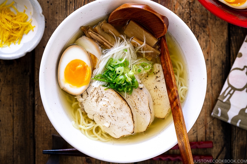 A white bowl containing Shio Ramen, topped with Chicken Chashu, Ramen Egg, bamboo shoot, white negi, and green onion.