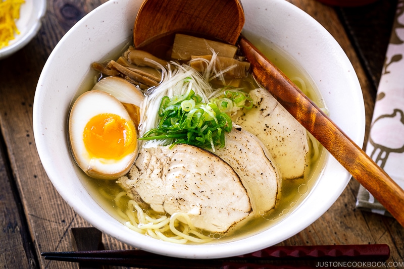 A white bowl containing Shio Ramen, topped with Chicken Chashu, Ramen Egg, bamboo shoot, white negi, and green onion.