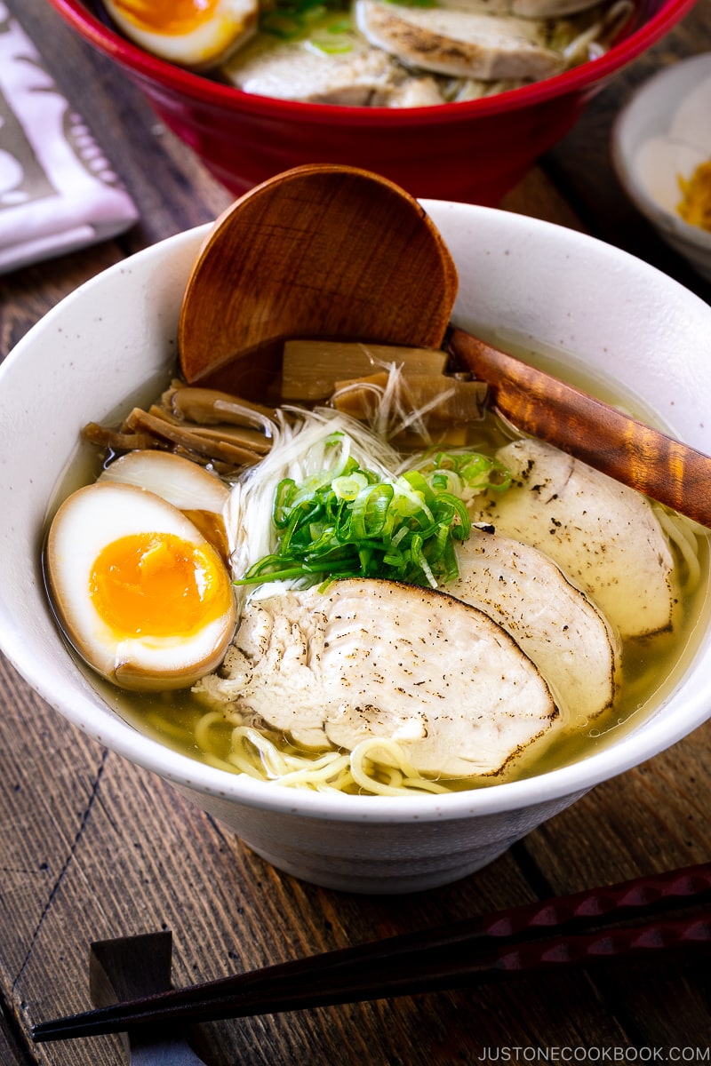 A white bowl containing Shio Ramen, topped with Chicken Chashu, Ramen Egg, bamboo shoot, white negi, and green onion.