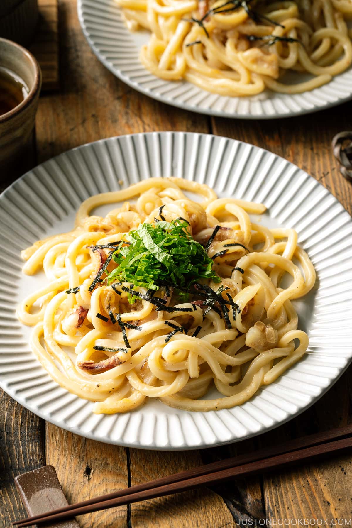 A white plate containing Mentaiko Udon topped with shredded nori and shiso leaves.