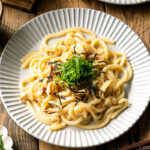 A white plate containing Mentaiko Udon topped with shredded nori and shiso leaves.