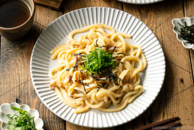 A white plate containing Mentaiko Udon topped with shredded nori and shiso leaves.