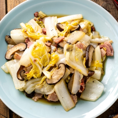 A round ceramic plate containing napa cabbage stir-fry with shiitake mushrooms and bacon seasoned lightly with soy sauce.