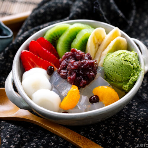 A bowl containing anmitsu, a traditional dessert with kanten jelly, fruits, mochi, green tea ice cream, and sweet red bean paste.