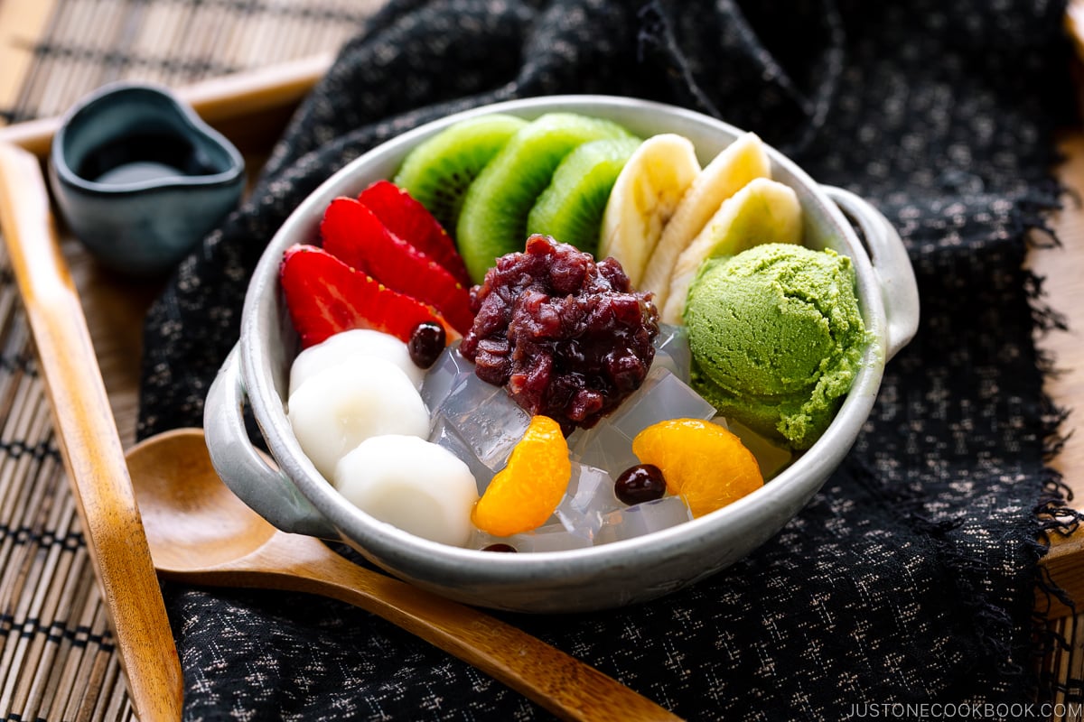 A bowl containing anmitsu, a traditional dessert with kanten jelly, fruits, mochi, green tea ice cream, and sweet red bean paste.