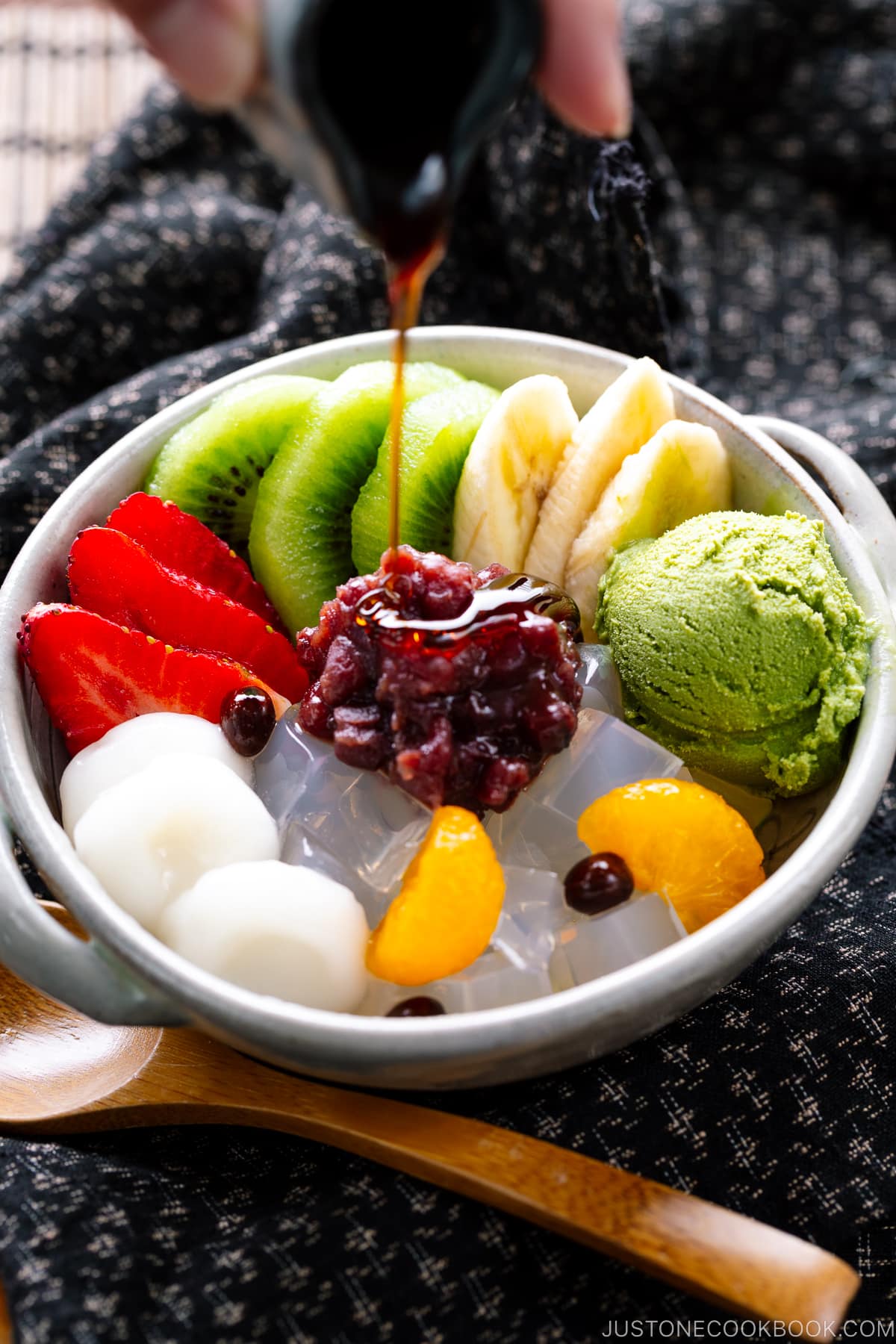 A bowl containing anmitsu, a traditional dessert with kanten jelly, fruits, mochi, green tea ice cream, and sweet red bean paste.