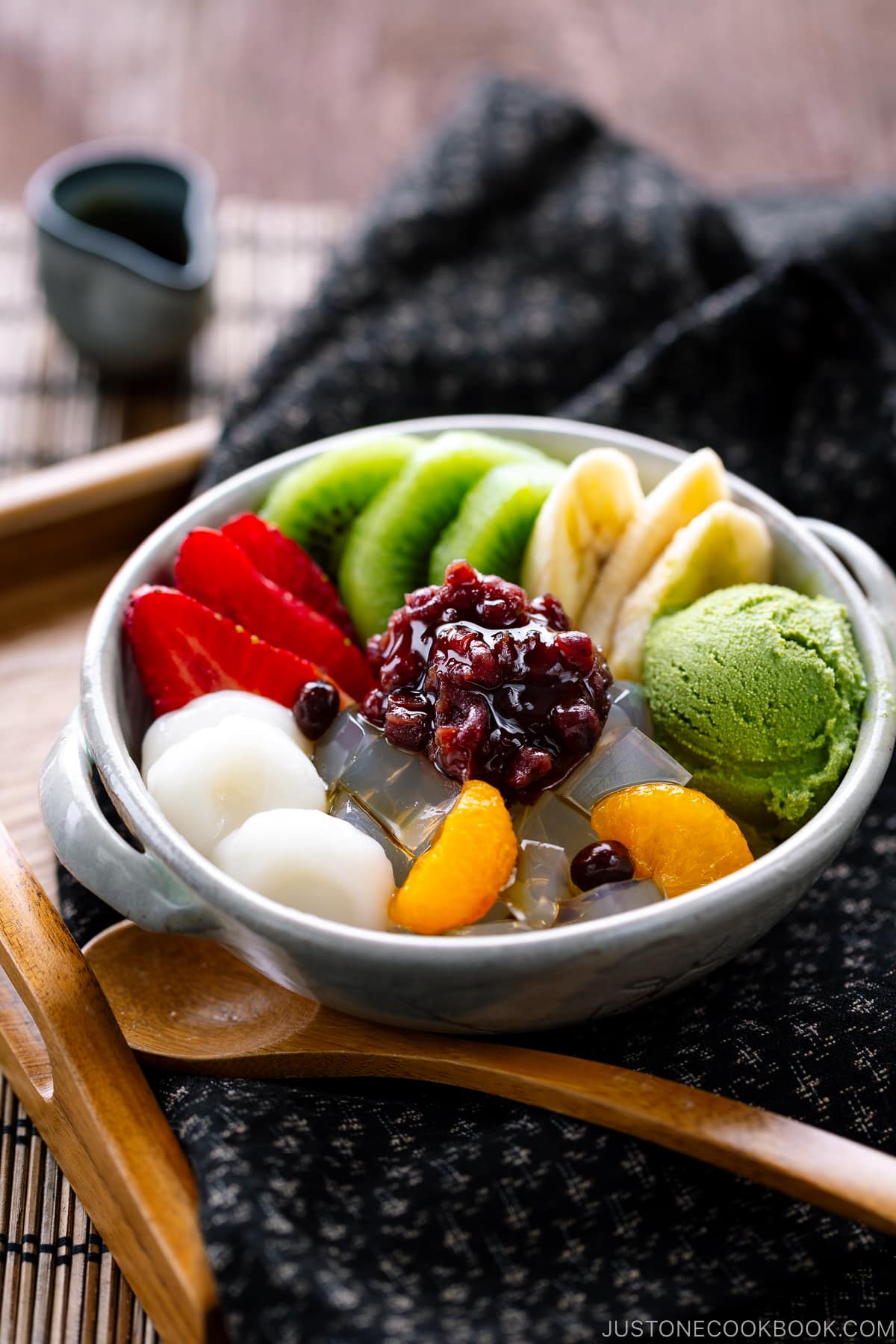 A bowl containing anmitsu, a traditional dessert with kanten jelly, fruits, mochi, green tea ice cream, and sweet red bean paste.