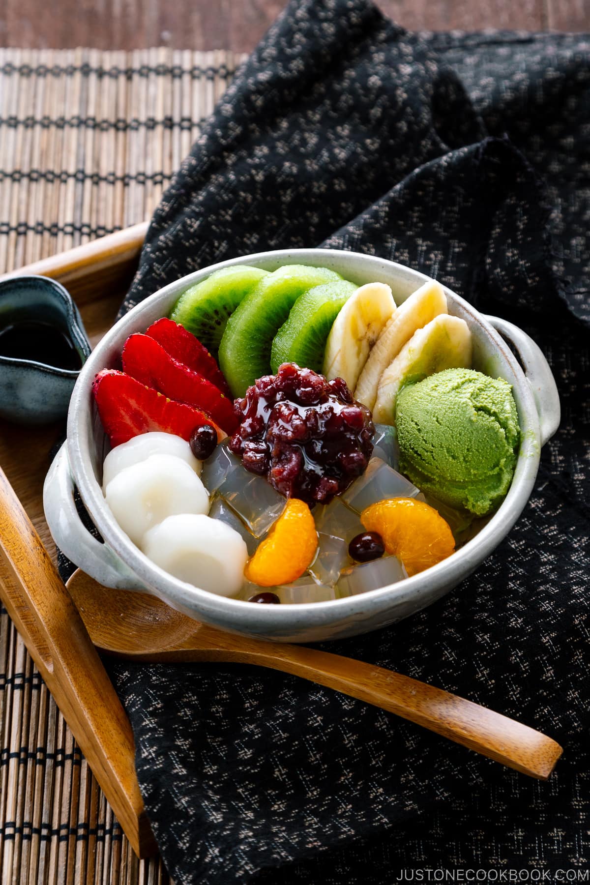 A bowl containing anmitsu, a traditional dessert with kanten jelly, fruits, mochi, green tea ice cream, and sweet red bean paste.