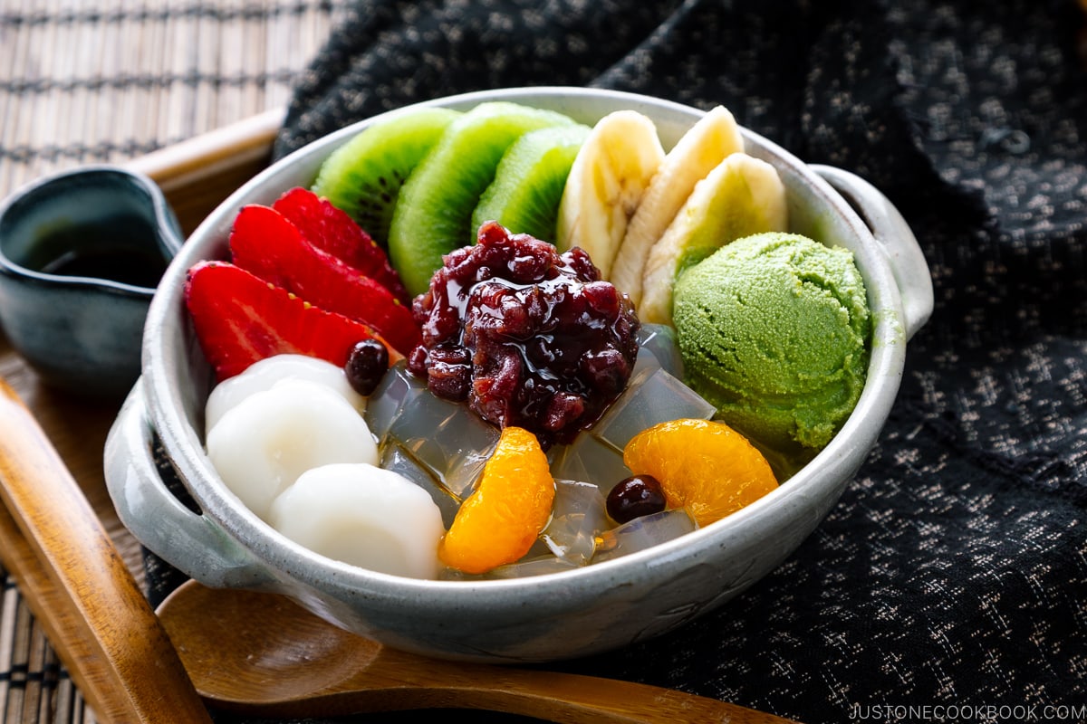 A bowl containing anmitsu, a traditional dessert with kanten jelly, fruits, mochi, green tea ice cream, and sweet red bean paste.