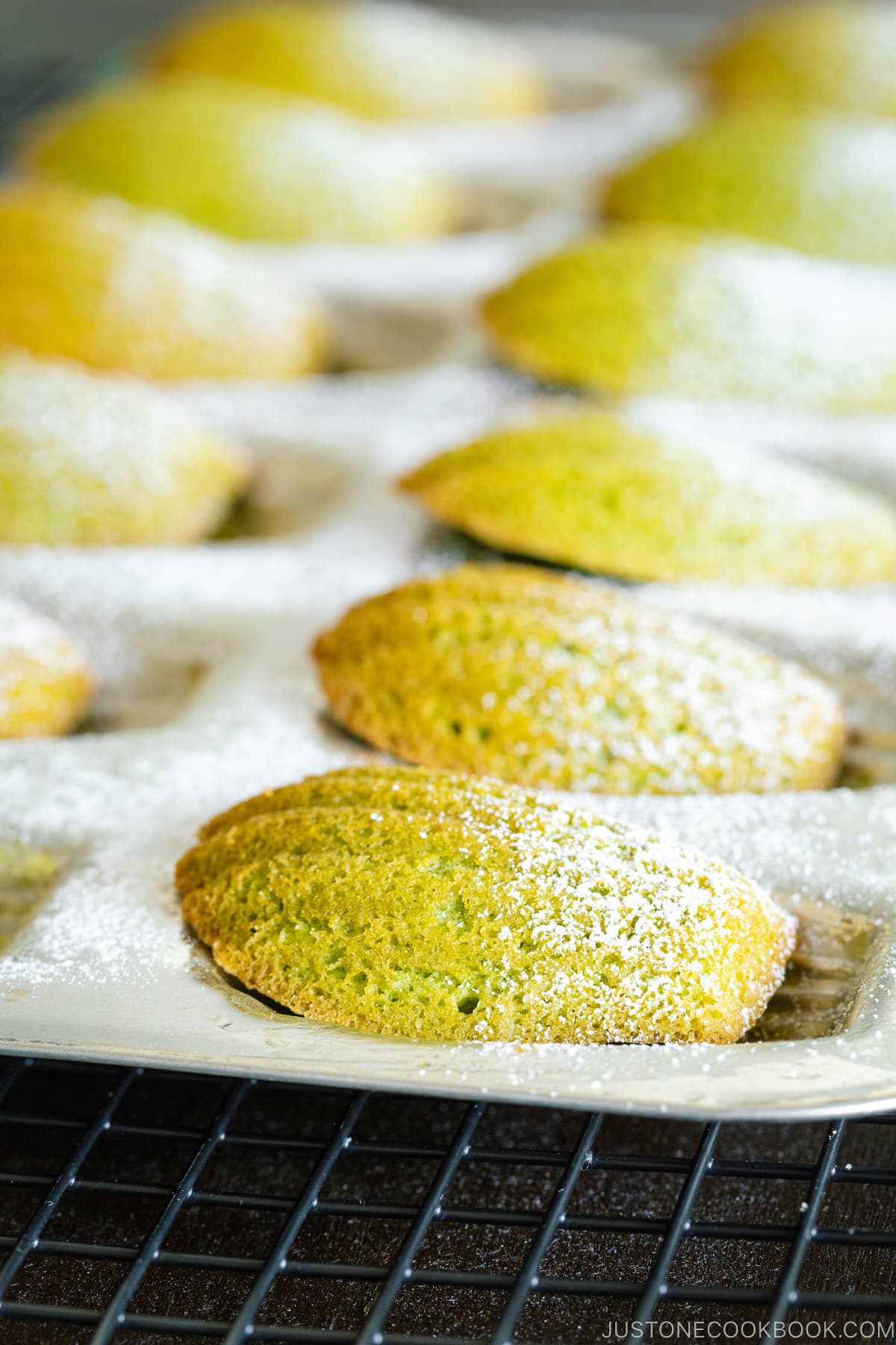 Un moule à madeleine contenant des madeleines au thé vert matcha saupoudrées de sucre en poudre.