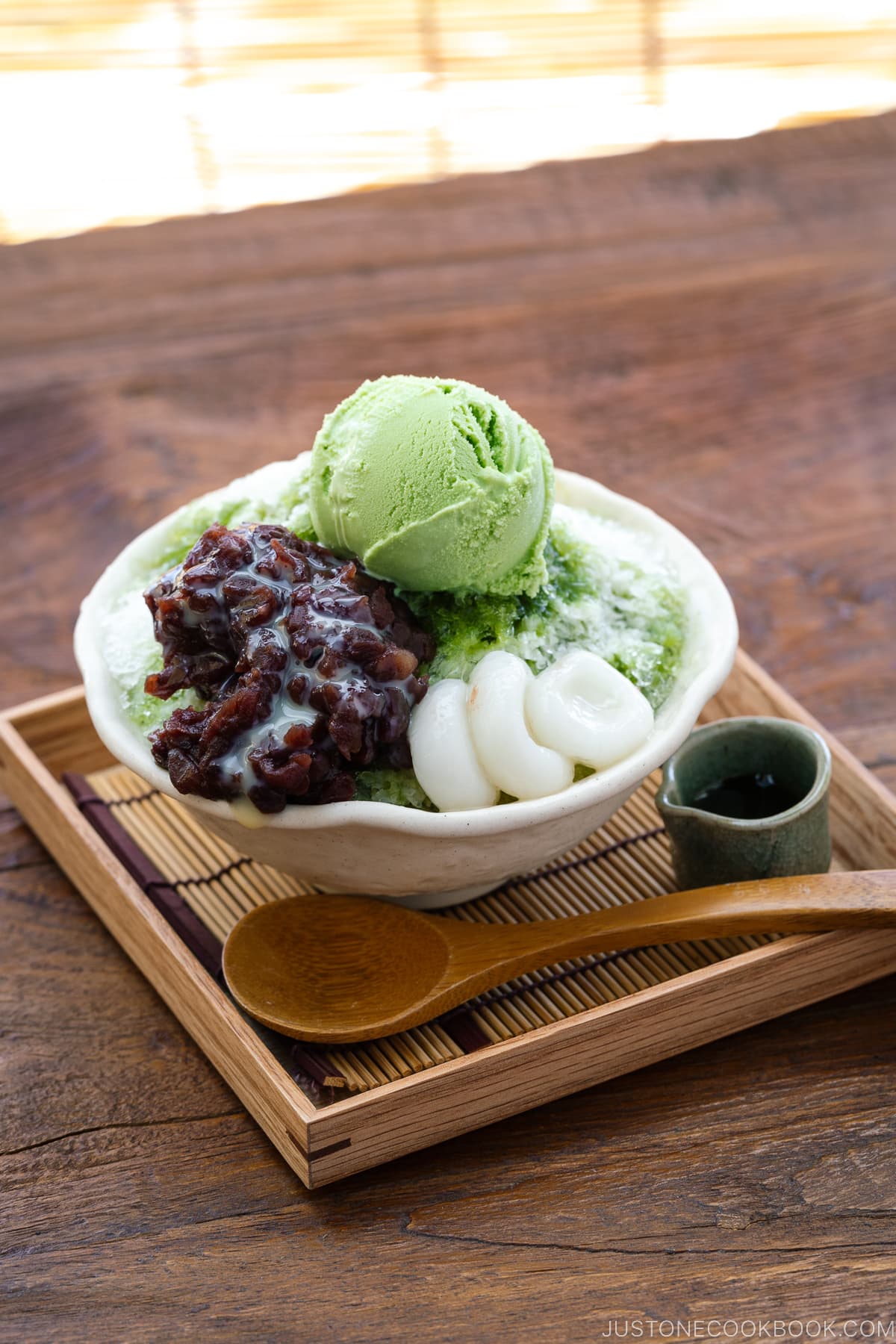 A bowl containing shaved ice, drizzled with matcha syrup and topped with red bean paste, shiratama dango, sweetened condensed milk, and green tea ice cream.