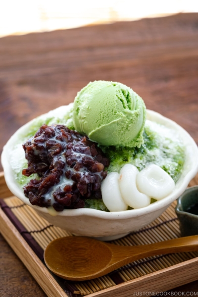 A bowl containing shaved ice, drizzled with matcha syrup and topped with red bean paste, shiratama dango, sweetened condensed milk, and green tea ice cream.