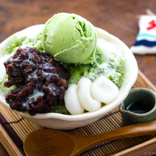 A bowl containing shaved ice, drizzled with matcha syrup and topped with red bean paste, shiratama dango, sweetened condensed milk, and green tea ice cream.